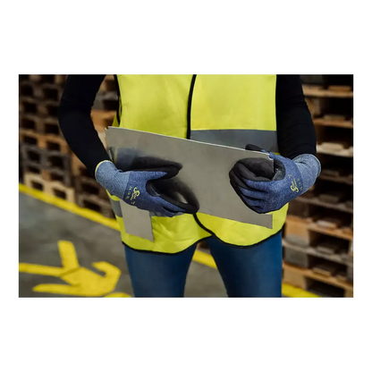 A person with a neon yellow safety vest and blue AMPri SolidSafety Cut cut-resistant gloves S - XXL from AMPri Handelsgesellschaft mbH carefully handles a large metal sheet in the warehouse. In the background, wooden pallets are stacked, yellow markings on the floor guide safe movements and emphasize occupational safety.