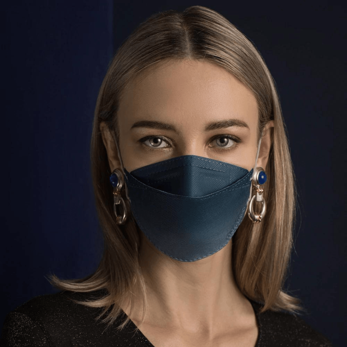 A person with long hair wears a blue Altruan designer face mask particle-filtering, providing effective protection according to FFP2 standard. She pairs it with large decorative earrings in front of a dark background and wears a black shirt.