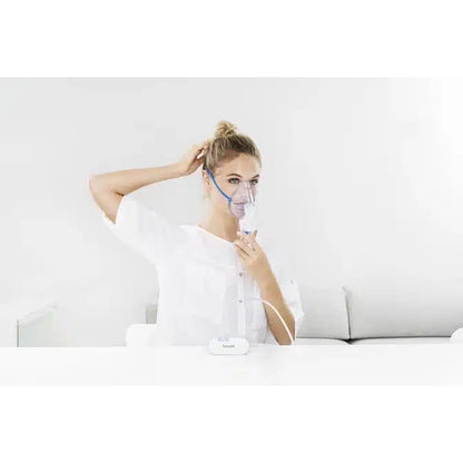 A person uses the discontinued product: Beurer small inhaler IH 60 from Beurer GmbH with a face mask at a white table and adjusts their hair. The minimal white background highlights this compact device for respiratory diseases.