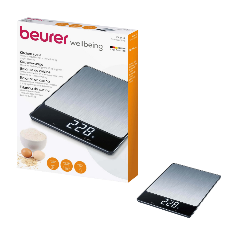 A digital kitchen scale in black and silver, the Beurer KS 34 XL stainless steel kitchen scale, is shown next to its packaging. On the packaging, it says "Beurer Wellbeing" and includes product features in several languages. This scale from Beurer GmbH features an LED display with "228" and a capacity of 15 kg, making it perfect for any kitchen task. Flour, sugar, and eggs are depicted in the background.