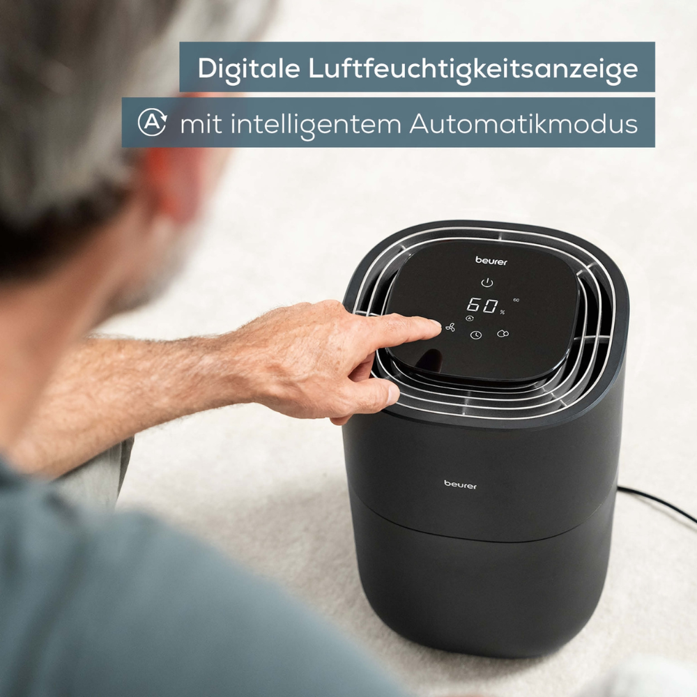 A person is seen interacting with a Beurer LB 200 humidifier from Beurer GmbH. The device's digital display shows a humidity of 50%. The accompanying text in German reads "Digital humidity display with intelligent automatic mode."