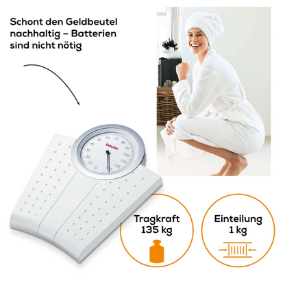 A woman in a white bathrobe with a towel on her head smiles while crouching next to a mechanical personal scale Beurer MS 50. The analog personal scale with analog weight display and non-slip surface features a text describing its benefits: "Economical for the wallet – no batteries required", with a load capacity of 135 kg and 1-kg increments.