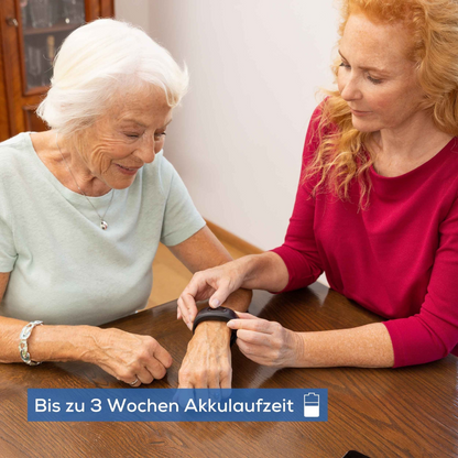An elderly woman sits at a table while a younger woman helps her put on a black Beurer emergency bracelet EC 70 SafePlus. Both are smiling. A German text overlay reads: "Up to 3 weeks of battery life."