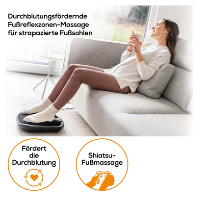 A woman relaxes on a gray couch and uses the Beurer FM 70 Shiatsu foot and back massager with optional heat function. She holds a remote control in her hands. She is wearing a white top and pink pants. Two symbols below describe the benefits: improved circulation and Shiatsu foot massage. German text with the brand name Beurer GmbH is displayed.