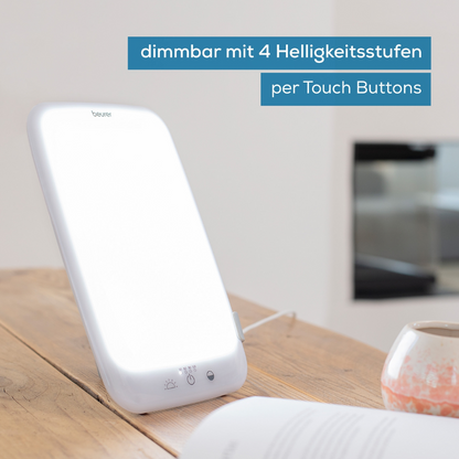 A rectangular Beurer daylight lamp TL 35 from Beurer GmbH stands on a table. The text on the image reads "dimmable with 4 brightness levels via touch buttons." In the background, a blurred living room setting can be seen. The light therapy lamp is illuminated and connected to a power source.