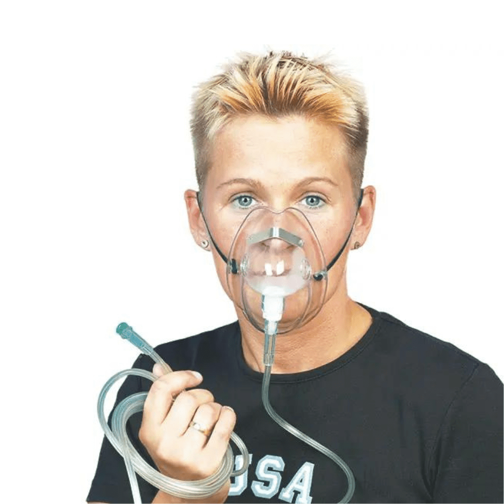 A person with short blonde hair is wearing the DCT oxygen mask for adults from Servoprax GmbH and is holding the attached O2 tube in their hand. They are wearing a black shirt with white lettering on a plain white background.