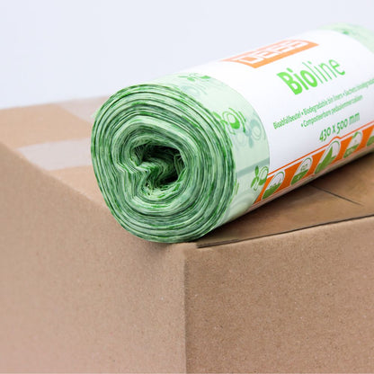 Eine Rolle grüner Biomüllbeutel wird auf einen Karton gelegt. Das Etikett auf der Rolle ist teilweise sichtbar und trägt den Aufdruck „EMIL DEISS KG (GmbH + Co.).“ Diese 100% kompostierbaren Beutel haben ein florales Design und sind 430 x 500 mm groß.