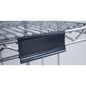Close-up of a Novocal label frame with paper tags (approx. 100 x 44 mm, 1 piece) attached to a metal wire shelf, with the black plastic label holder on the front edge; the background is blurred.