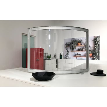 A Novocal hygiene protection wall, curved with a pass-through (1,200 x 600 mm), stands on a white counter with a geometric black bowl and a small black object in front of it. Its reflection shows a red refrigerator, green bottles, and shelves with potted plants.