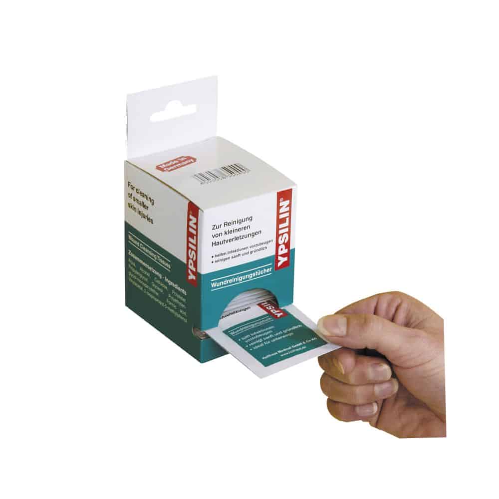 A hand pulls a Holthaus Medical YPSILIN® wound cleansing cloth from a box dispenser. The packaging has German text, and the box is white and green, with clearly visible product information and the brand name of Holthaus Medical GmbH & Co. KG.