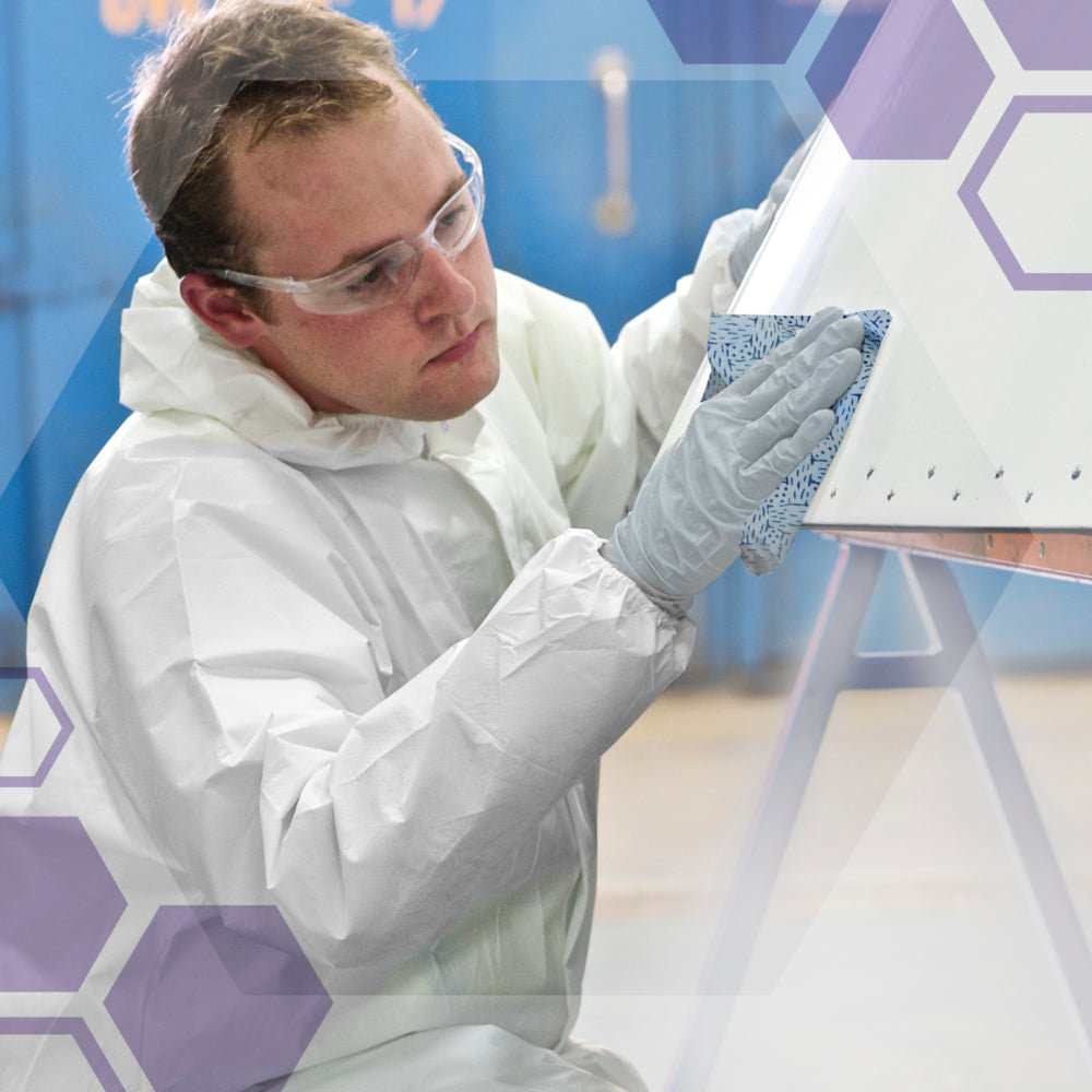 A person wearing safety glasses, gloves, and a white suit uses Kimberly-Clark GmbH Kimtech® process wipes (BRAG™ Box, Blue, 160 wipes) to clean a white object indoors, with purple hexagons indicating innovative industrial cleaning.