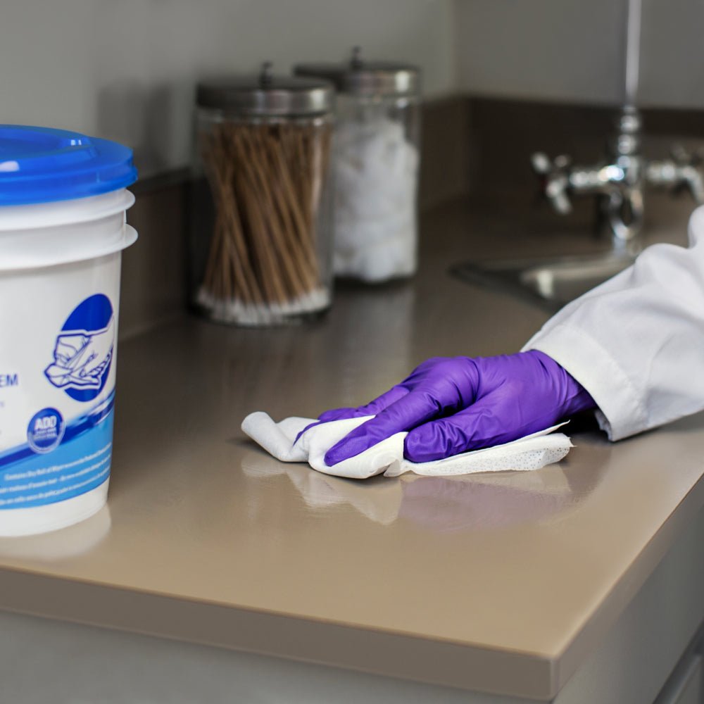 A person with purple gloves wipes a beige countertop near the sink with WypAll® Wettask™ wipes from Kimberly-Clark GmbH (6 rolls x 90 sheets), while jars with cotton swabs and balls are visible in the background.