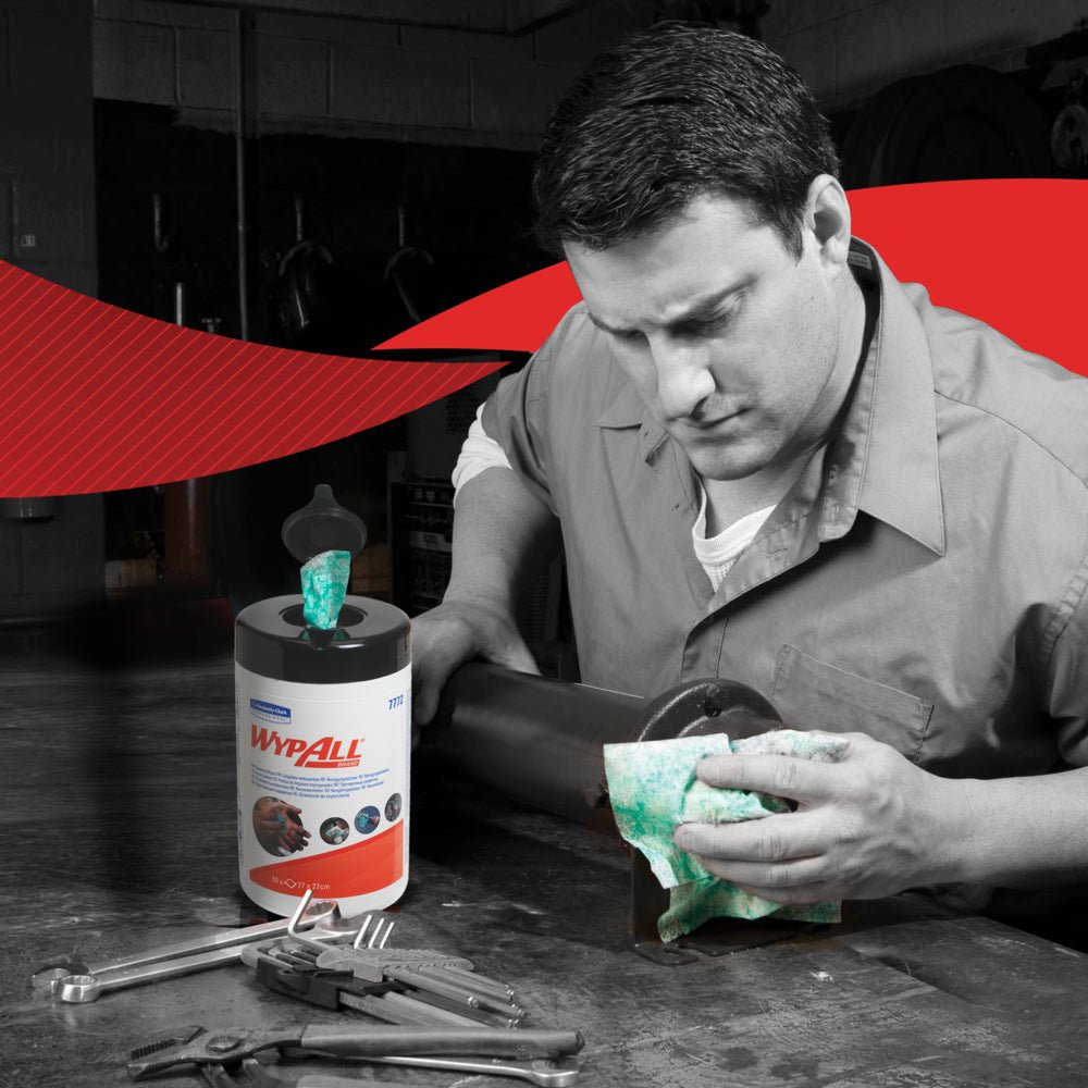 A man in a work shirt wipes a metal object with a green WypAll® wipe from Kimberly-Clark GmbH in a workshop. On the table are the WypAll® wipes - Green (6 Tubs x 50 Sheets | Carton) and hand tools. The background is predominantly black and white with red accents.