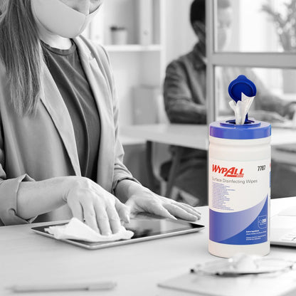 A masked person disinfects a tablet at a desk with WypAll® surface disinfection wipes (dispenser bucket, 200 sheets) from Kimberly-Clark GmbH, with the tray visible, while another person works at a computer in the background.