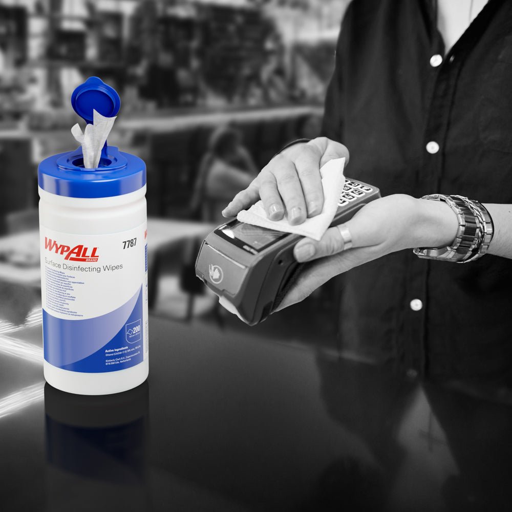 Someone uses a WypAll® surface disinfection wipe (Kimberly-Clark GmbH) from an open dispenser tray to disinfect a card terminal. The white dispenser bucket (200 wipes) is on the counter, the background is blurred.