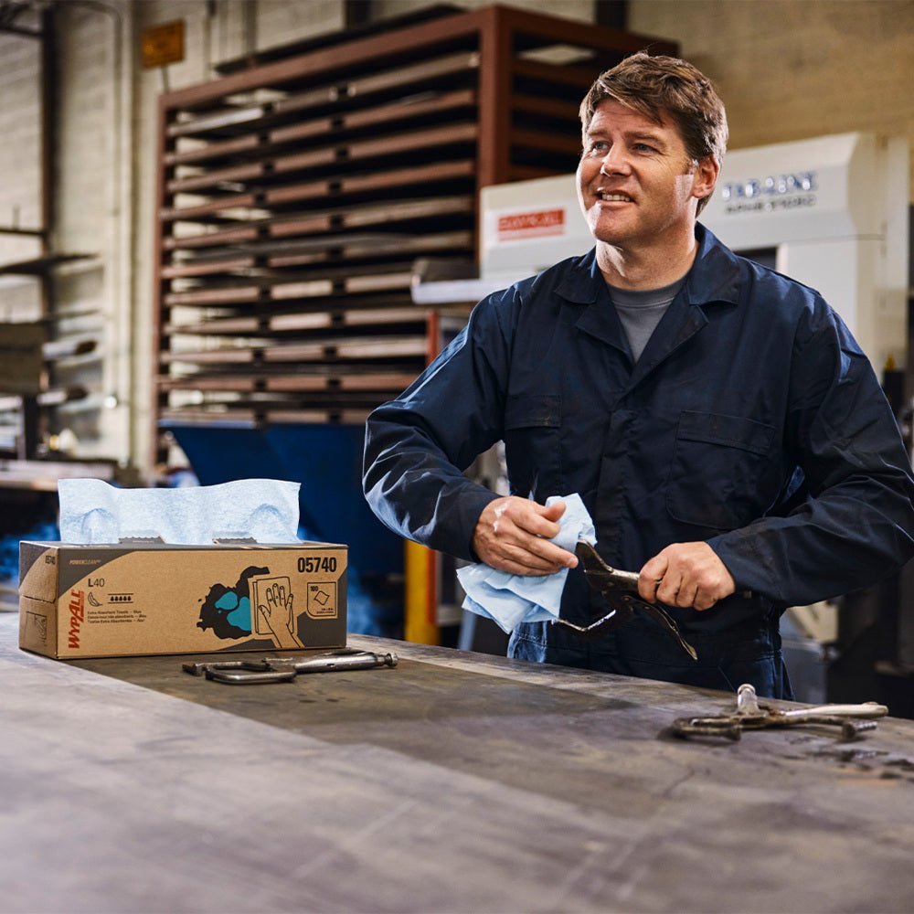 A man in a navy blue overall wipes a tool with a blue WypAll® X80 PowerClean cloth from a Steel Blue pull-up box (Kimberly-Clark GmbH) while standing at a workbench with metal tools and boxes of WypAll cloths in an industrial workshop.
