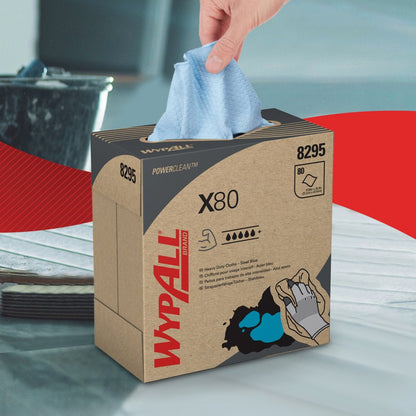 A hand pulls a steel blue WypAll® X80 PowerClean cleaning cloth from a pull-up box from Kimberly-Clark GmbH, clearly showing product details. In the blurred background, a bucket and a red-white area can be seen.