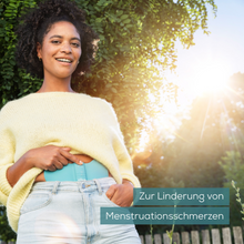 A person in a yellow sweater and jeans stands outdoors holding a Beurer EM 55 Menstrual Relax+ Gel-Pad from Beurer GmbH in their hand. Green is visible in the background, with sunlight filtering through. The text reads "For the relief of menstrual pain".