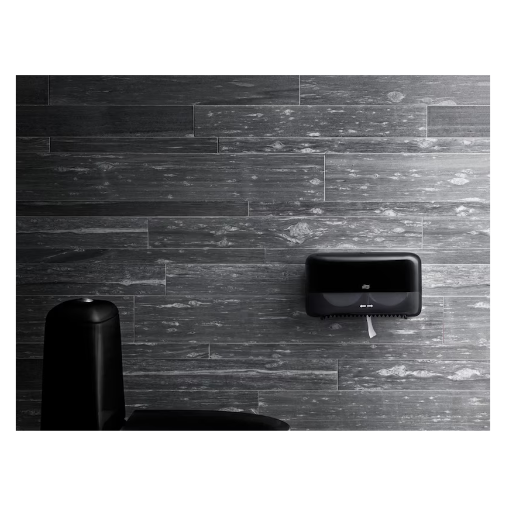 A modern bathroom with dark gray textured walls presents a high-quality wall-mounted Tork paper towel dispenser in black and partially reveals an elegant black trash can in the foreground, while maintaining an eco-friendly design.