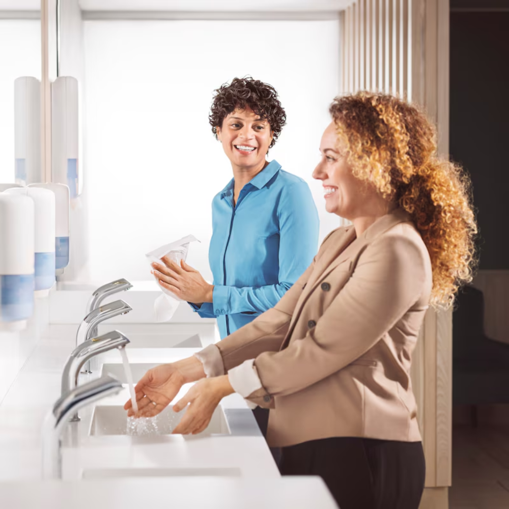 Two women are standing at the sinks of a public restroom. One is washing her hands with Tork 520201 Pure Hand Foam Soap Premium S4 (1000 ml) from TORK – Essity Professional Hygiene Germany GmbH, the other is smiling and holding paper towels in her hand. Soap dispensers are on the washstand.