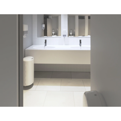 A clean, modern public restroom features a long white countertop with two sinks. Large mirrors are mounted above the sinks. Tork 553100 Mini Dispensers for Zigzag Hand Towels Elevation H3 by Essity Professional Hygiene Germany GmbH are mounted on the wall, and to the left of them is a trash can. The floor is tiled with light tiles and features a relief design.