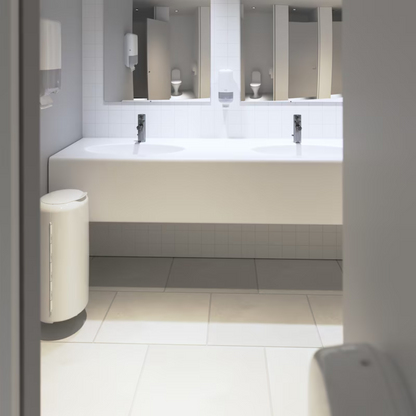 A clean, modern bathroom presents an Elevation design with a long white countertop featuring two sinks with chrome fixtures. The walls are adorned with white tiles and mirrors are mounted above the sinks. Tork 561500 dispensers for soap and hand sanitizer Elevation S4 by TORK – Essity Professional Hygiene Germany GmbH are elegantly mounted on the wall. An elegant white trash bin stands on the floor.