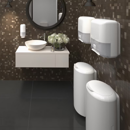 A modern bathroom with dark mosaic tiles features a round mirror above a white countertop sink. To the right, two Tork Matic® 551100 sensor dispensers for roll towels Elevation H1 by TORK – Essity Professional Hygiene Germany GmbH provide touch-free hand drying and single sheet dispensing. Below, two white waste bins and a bouquet of white flowers add a decorative touch to the countertop.