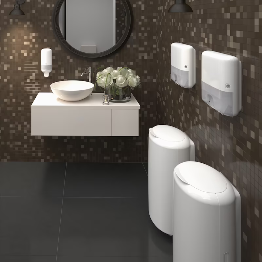 A modern public restroom with a wall of brown mosaic tiles features a round mirror, a wall-mounted sink with a white basin, soap dispenser, Tork Xpress® 552100 Mini Dispenser for Multifold Hand Towels Elevation H2 by TORK – Essity Professional Hygiene Germany GmbH, and two white cylindrical trash bins. A bouquet of white flowers decorates the sink area.