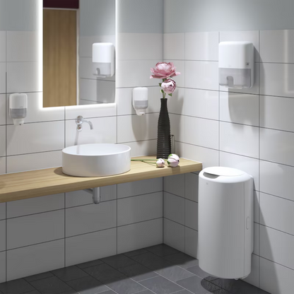 A modern bathroom with a round white sink on a wooden countertop, enhanced by a wall mirror with lighting. The Tork Xpress® 552100 Mini Dispenser for Multifold Hand Towels Elevation H2 by TORK – Essity Professional Hygiene Germany GmbH adds elegance to the white-tiled walls, along with a black vase full of pink flowers. A white trash bin stands on the gray-tiled floor.