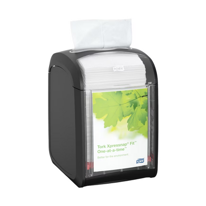 The Tork Xpressnap Fit® 272900 table dispenser for napkins by TORK – Essity Professional Hygiene Germany GmbH elegantly holds white napkins. The front label adorned with green leaves proudly states "Tork Xpressnap Fit One-at-a-time," emphasizing reduced napkin consumption and commitment to the environment.