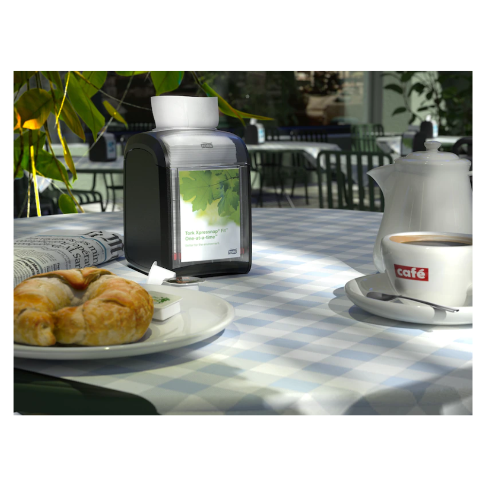A table in a café, adorned with a blue and white checkered tablecloth, is elegantly set for one person. On the table are a croissant, a cup of coffee in a "Café" cup, a teapot, and a newspaper. The Tork Xpressnap Fit® 272900 table dispenser for napkins by TORK – Essity Professional Hygiene Germany GmbH sits prettily beside it. Sunlight filters through the nearby plants.