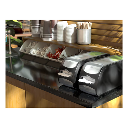 A coffee station is equipped with a Tork Xpressnap Fit® 272901 countertop napkin dispenser from TORK - Essity Professional Hygiene Germany GmbH, which has two napkin dispensers. Additionally, neatly stacked paper cups, lids, stir sticks, and condiments like sugar and cream packets are arranged in compartments on an elegant black counter.