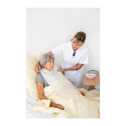 A nurse adjusts the blanket of an elderly woman who sits worriedly in bed. The nurse is wearing a white uniform with a Vala® Fit armband from Paul Hartmann AG and the room appears bright and clean.