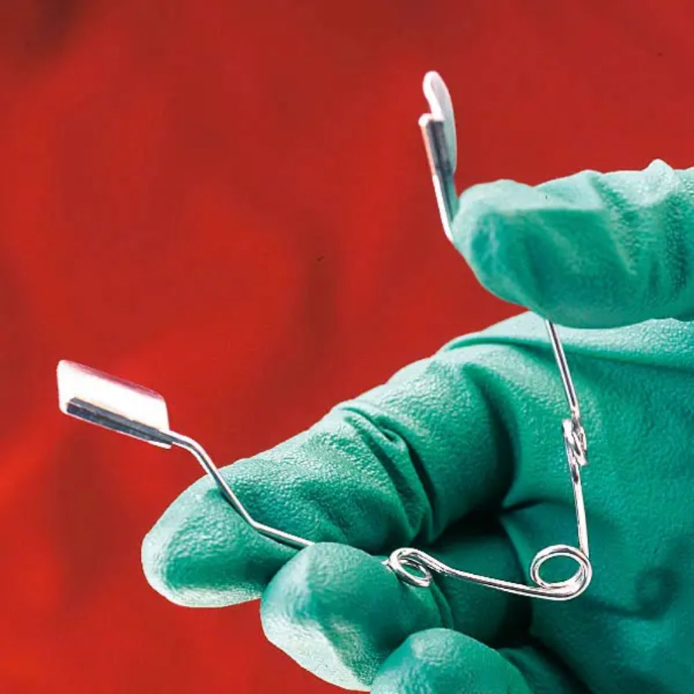 A person with a green glove holds an Altruan rabbit cheek retractor, a metal dental retractor with flat, angled ends and a wire-like structure, in front of a red background.