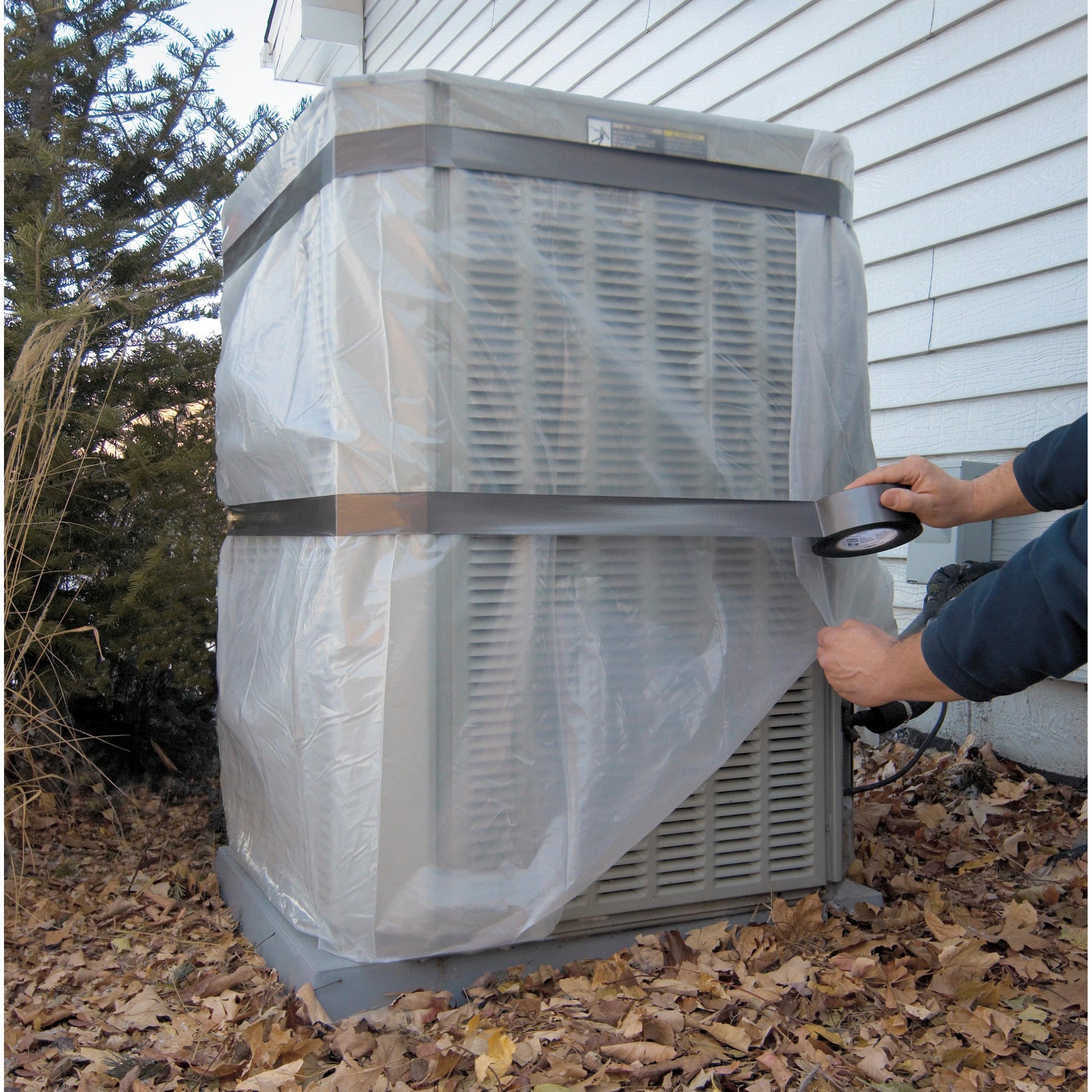 Jemand sichert ein großes Außenklimagerät neben einem Haus mit dem 3M Scotch® Extremium™ All Weather Hochleistungs-Klebeband (27 m x 48 mm) und Plastikfolie inmitten von trockenen Blättern und Pflanzen.