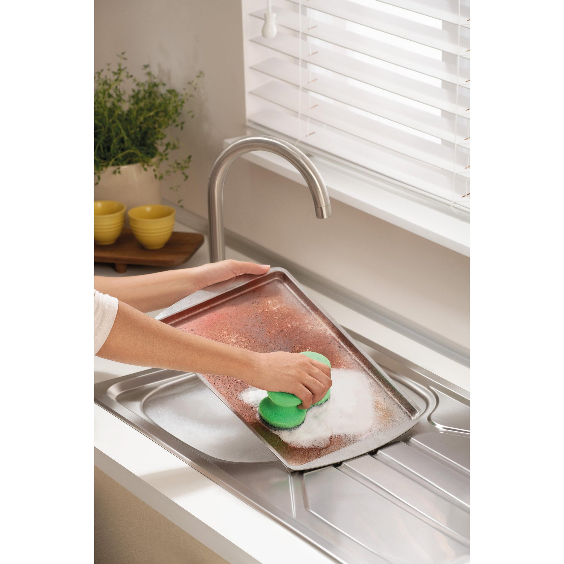 Jemand schrubbt ein großes, verschmutztes Backblech mit dem Scotch-Brite® Classic Reinigungsschwamm von 3M Deutschland GmbH unter einem Wasserhahn in einer hellen Küche mit weißen Jalousien, einer Pflanze und gestapelten gelben Schalen im Hintergrund.