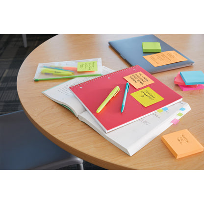 A round wooden table with a closed laptop, notebooks, pens in a plastic bag, highlighters, reminder notes, and a stack of 3M Deutschland GmbH Post-it® Super Sticky Notes Cube, Yellow (76 mm x 76 mm). A chair is partially visible.