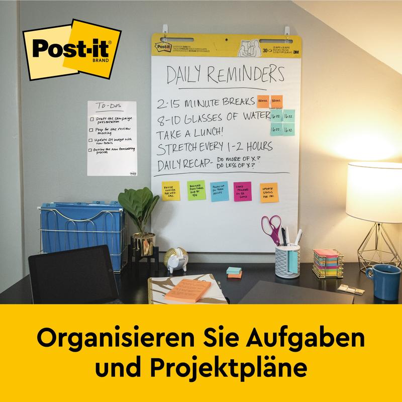 A desk with office supplies, lamp, and chair stands in front of a wall-mounted 3M Post-it® Super Sticky self-adhesive Meeting Chart (63.5x76.2 cm, 30 sheets) that shows daily reminders and notes. On a yellow banner below it says "Organize tasks and project plans."