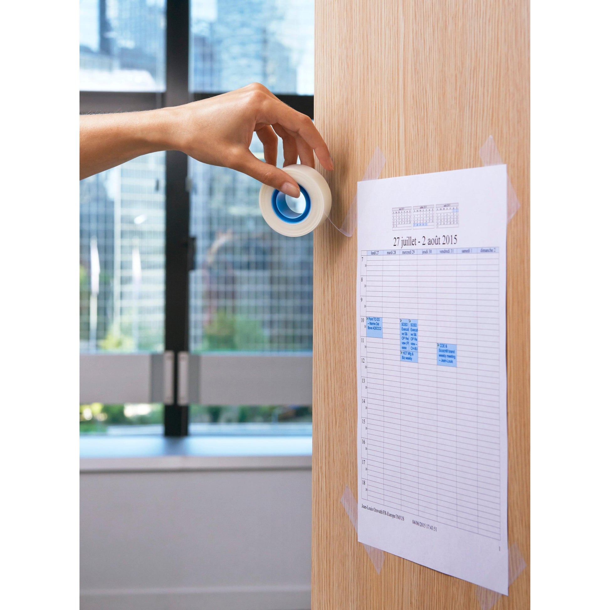 Eine Hand hält eine Rolle Scotch® unsichtbares Klebeband, ablösbar (19 mm x 33 m, 1 Stück) von 3M Deutschland GmbH neben einem Wochenplan, der auf einer Holzfläche befestigt ist. Im Hintergrund sieht man ein Fenster mit Bäumen und Gebäuden.