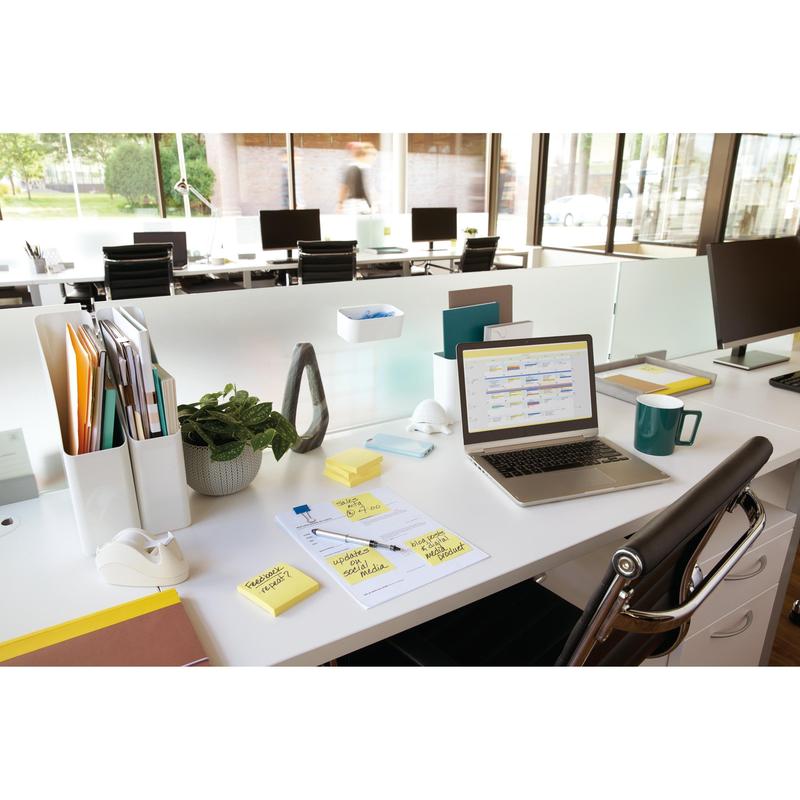 A modern office workspace with a laptop, cup, notepads, pen, plant, tape dispenser, folders, and 3M Deutschland GmbH Post-it® Notes (Yellow, 76 mm x 127 mm) on a white desk. Large windows let in natural light; empty desks are visible behind.
