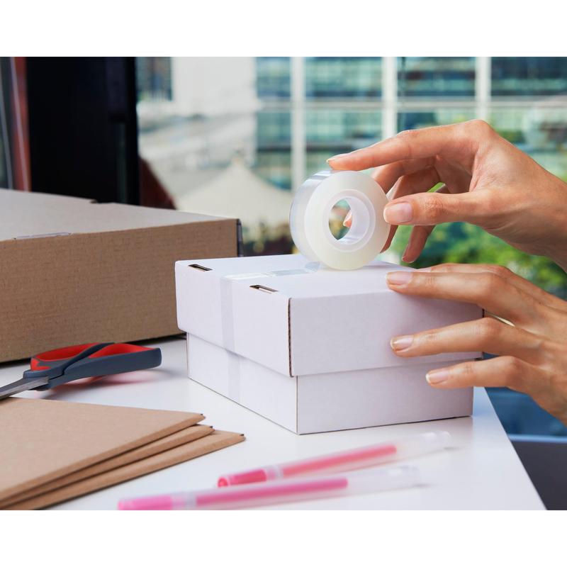 Someone seals a small white cardboard box with Scotch® Crystal Tape, transparent, 1 roll, 19 mm x 33 m by 3M Deutschland GmbH. A second box, scissors, brown cardboard sheets, and two pink pens are nearby on a light table.