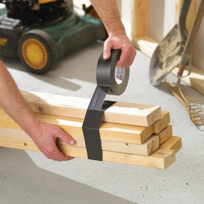 In einer Garage wickelt jemand das Scotch® Extremium™ Ultra Hochleistungs-Klebeband DT17 (25 m x 48 mm) der 3M Deutschland GmbH um Holzbretter, während im Hintergrund ein Rasenmäher, eine Harke und eine Schaufel zu sehen sind.