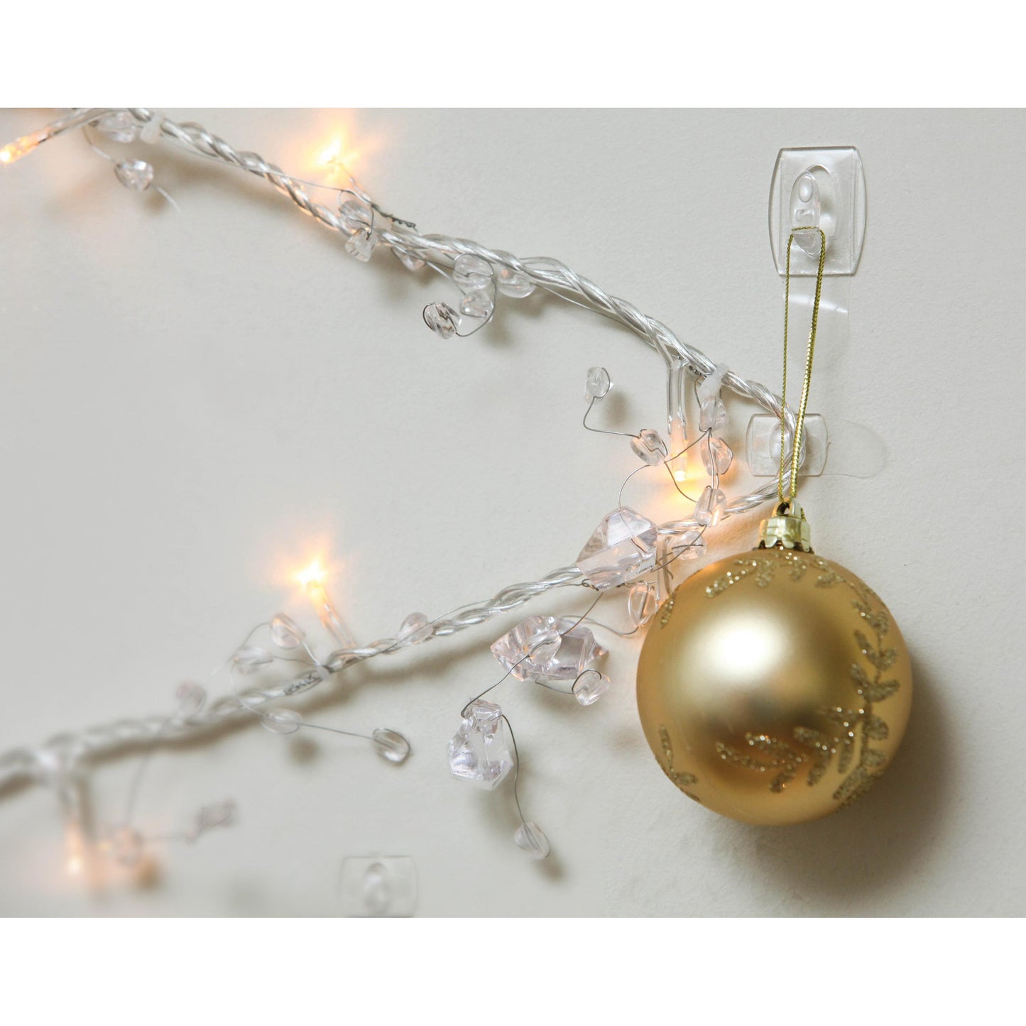 Ein goldenes Weihnachtsornament hängt an einem Command™ Mini-Haken, transparent (17006CLRFGN) von 3M Deutschland GmbH, an einer weißen Wand neben leuchtenden weißen Weihnachtslichtern - perfekte Aufbewahrung ohne Bohren!.