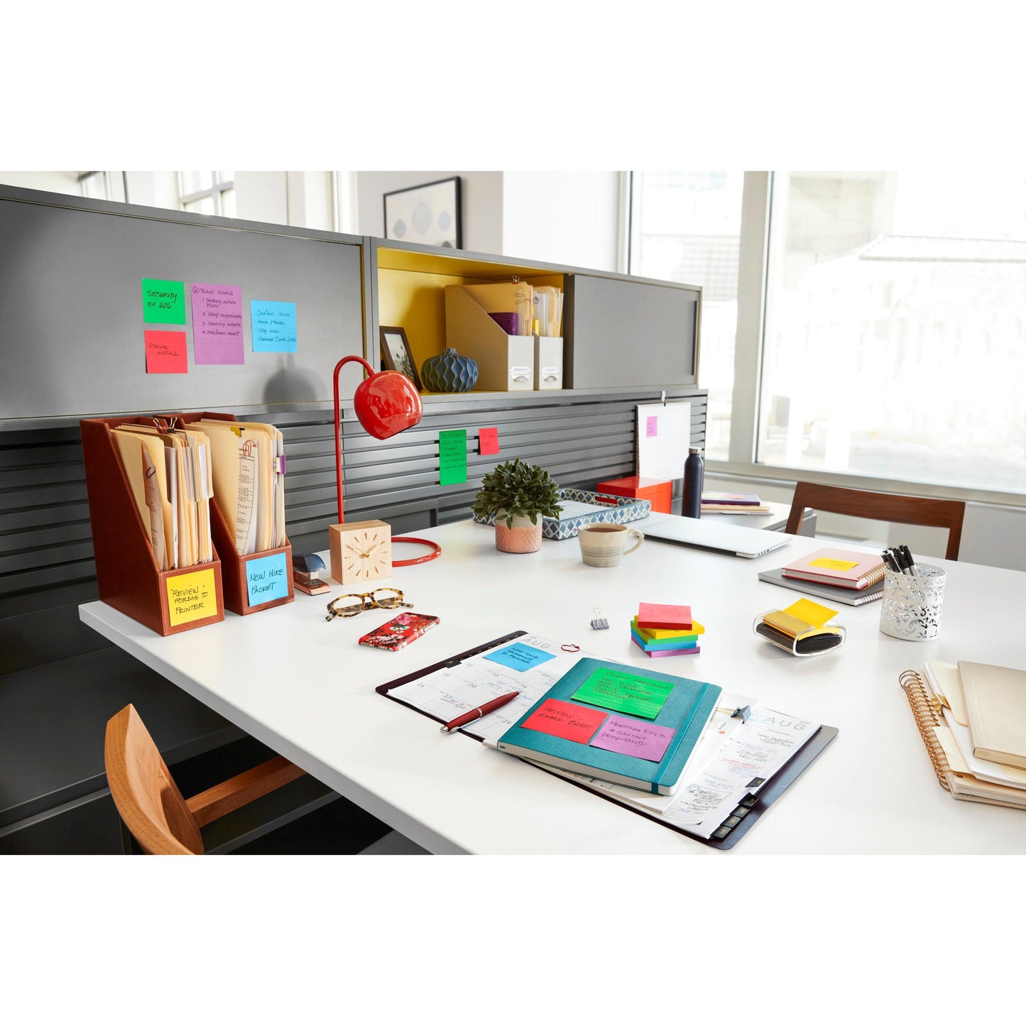 Ein aufgeräumtes Büro mit Post-it® Super Sticky Notes (47,6 mm x 47,6 mm, 90 Blatt/Block, 100 % PEFC) der 3M Deutschland GmbH, Ordnern, Notizbüchern, einer roten Lampe, einer Topfpflanze und anderen Utensilien, geordnet auf einem weißen Schreibtisch und grauen Regalen.
