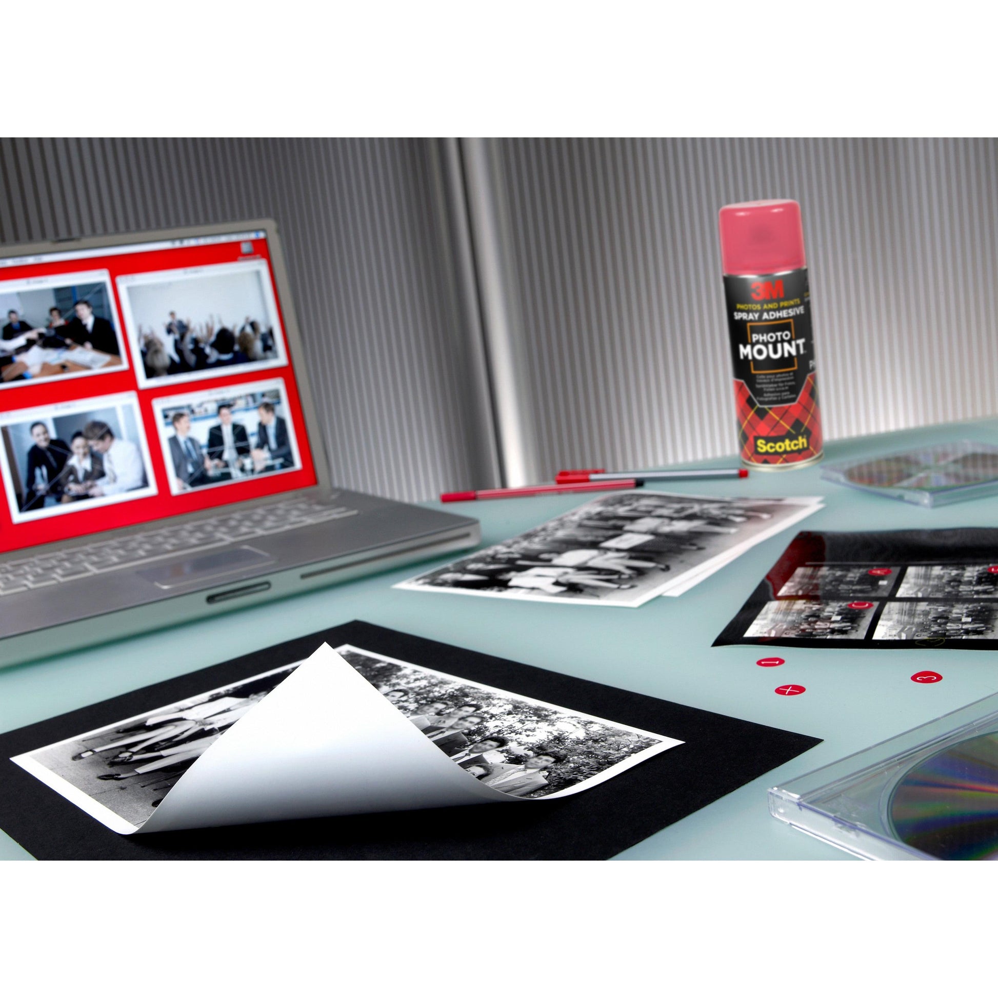 Auf einem Schreibtisch stehen Schwarz-Weiß-Fotos, ein Laptop mit Foto-Miniaturansichten, ein roter Stift, CDs, kleine rote Aufkleber und eine Dose 3M™ Scotch® PhotoMount™ Klebespray (400 ml) der 3M Deutschland GmbH auf einer hellen Fläche.