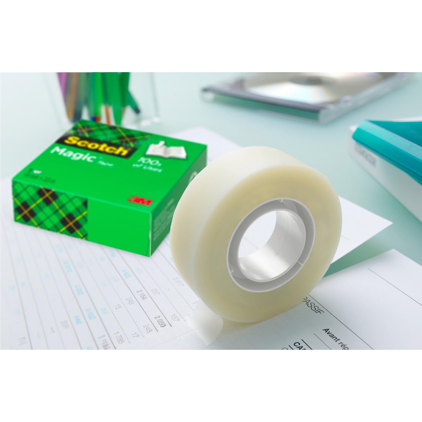 Eine Rolle 3M Deutschland GmbH Scotch® Magic™ Unsichtbares Klebeband, Vorteilspack 8 Rollen (19 mm x 33 m), liegt neben der grünen Schachtel mit auf Papier abgerolltem Klebeband; im Hintergrund sind unscharfe Büroartikel zu sehen.