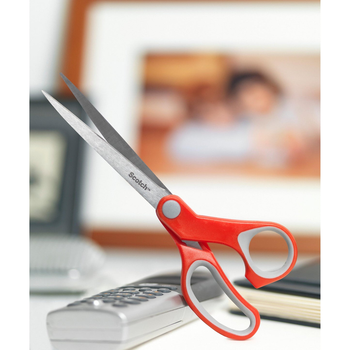 A Scotch™ Comfort Scissors, red by 3M Deutschland GmbH, rests on a corded phone, with blurred framed photos and books in the background.